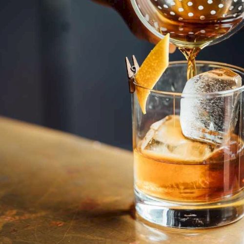 A cocktail being poured into a glass with ice, garnished with a twist of citrus peel, on a bar counter.
