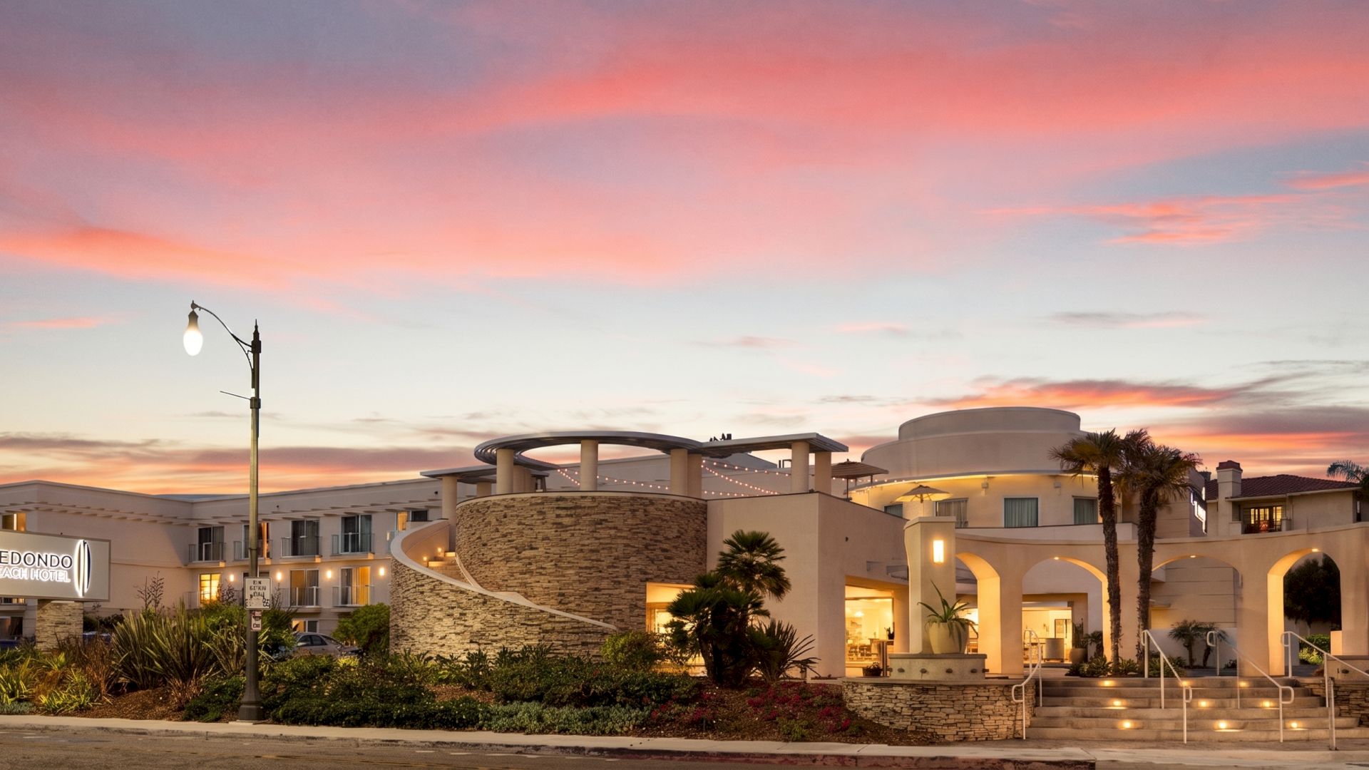 A modern, airy building with curved white walls sits by a palm-lined street at sunset, lit lamps glowing as pink and orange clouds fill the sky.