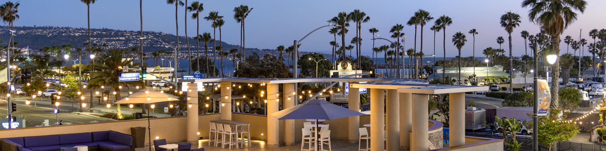 A rooftop patio at dusk with string lights, chairs and umbrellas, tall palm trees, and a view of the ocean and shoreline.