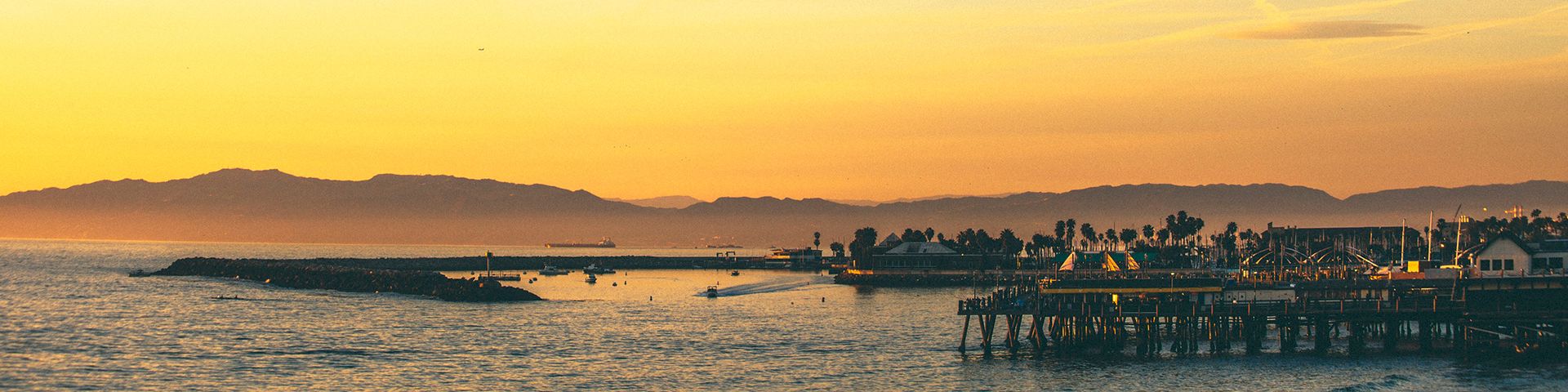 Sunset over a calm harbor with a pier, distant hills, and silhouettes of boats and palm trees along the shoreline.