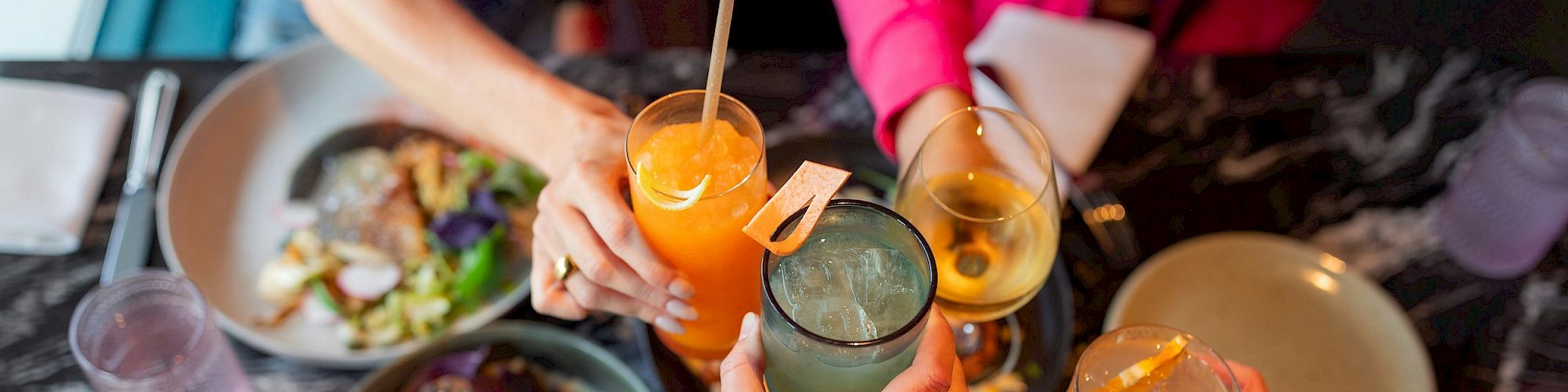 A group of friends cheers with colorful cocktails over a spread of bowls and plates at a social meal.
