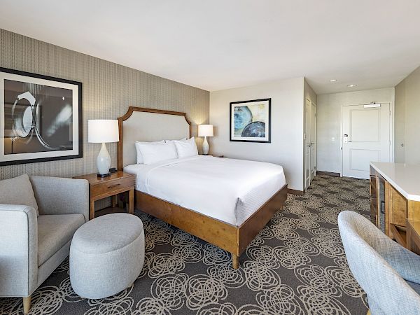 Bright hotel room with a neatly made bed, bedside lamps, framed artwork, a gray sofa, and a carpeted hallway view.