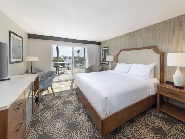 A modern hotel bedroom with a made bed, desk and TV, lamps, framed art, and large windows opening to outdoors.