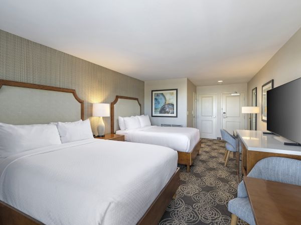 A bright hotel room with two beds, bedside lamps, framed art, a large TV, and a patterned carpet.