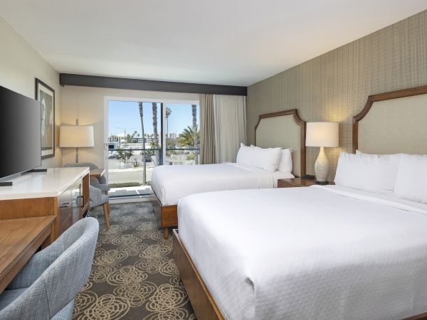 A modern hotel room with two beds, bedside lamps, a TV on a dresser, and large windows overlooking palm trees.
