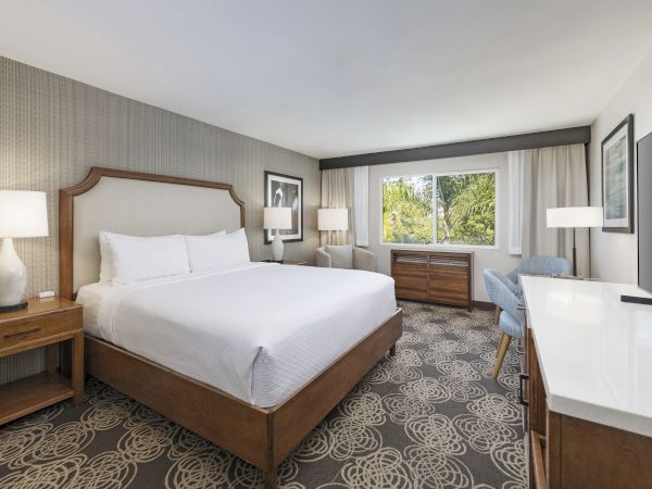 A modern hotel room with a large bed, lamps, curtains, a window view of palm trees, and a desk with chair.