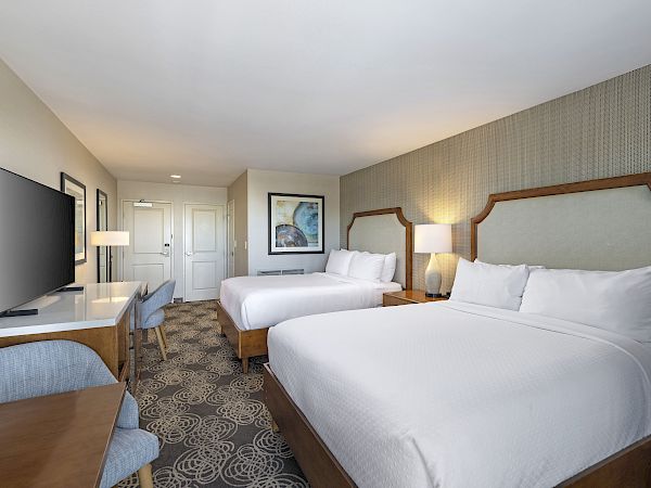 A tidy hotel room with two beds, a desk and TV, bedside lamps, and framed art on textured walls, under a ceiling light.