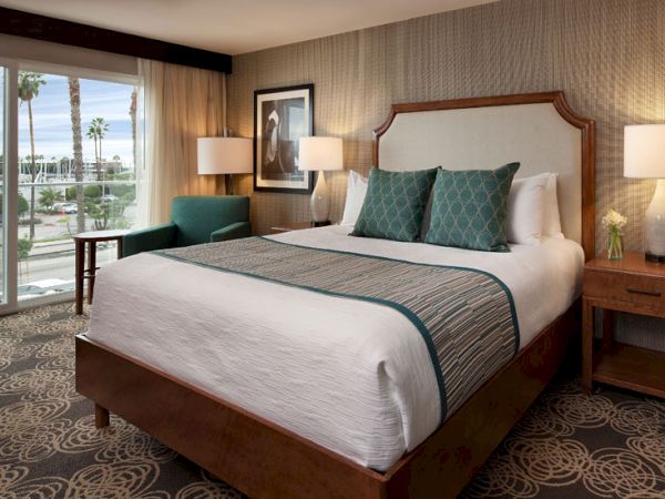 A cozy hotel room with a large bed, teal accents, two bedside lamps, a view of palm trees and the ocean outside, and warm lighting.