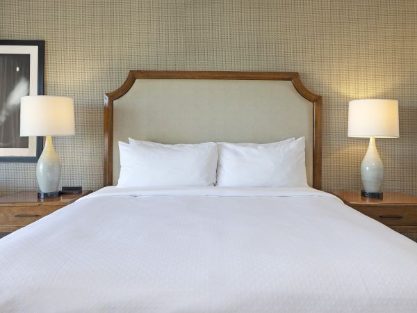 A neatly made hotel bed with white pillows and a wood headboard, flanked by two lit bedside lamps.