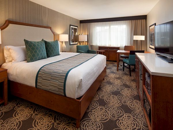 A modern hotel room with a king bed, teal accents, two nightstands, work desk, and a large window overlooking the city, all warmly lit.