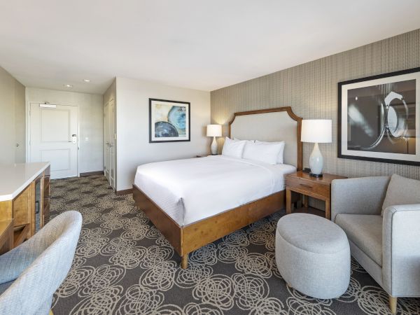 Modern hotel bedroom with a large bed, lamps, sofa, wall art, patterned carpet, and a kitchenette/desk area.