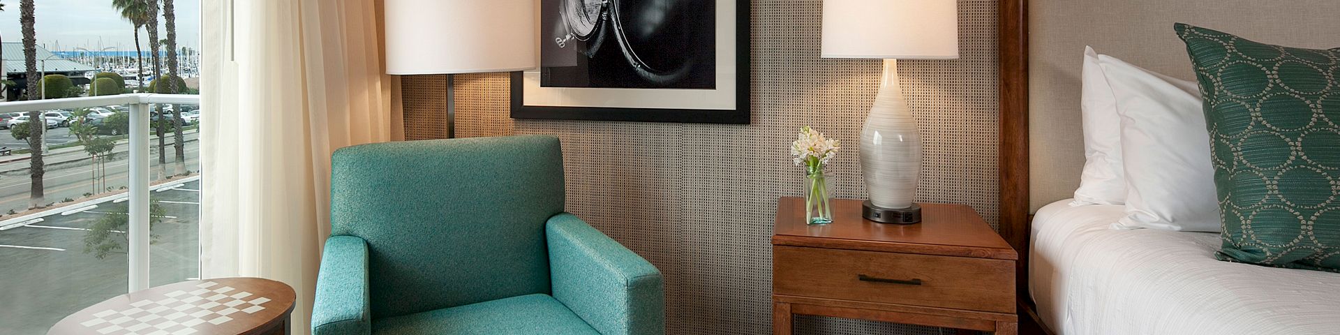 A stylish hotel room with a teal armchair, wooden nightstand, white lamps, framed art, and a bed with green patterned pillow, near a window.