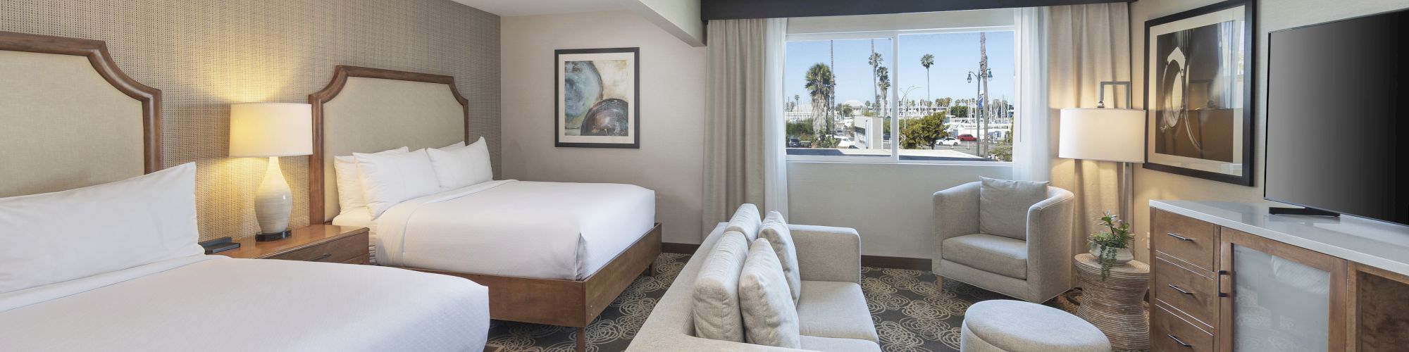 Modern hotel room with two beds, chairs, lamps, TV console, and a large window showing palm trees outside.