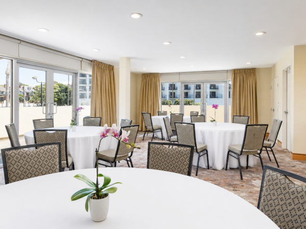A bright hotel conference room with round and banquet tables, black chairs, beige curtains, and white tablecloths with orchids.
