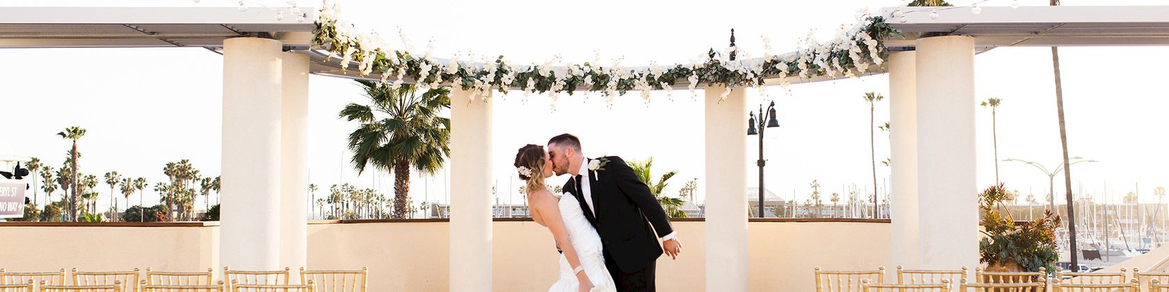 A bride and groom kiss at an outdoor wedding altar, surrounded by gold chairs and hanging garlands, on a sunny terrace.