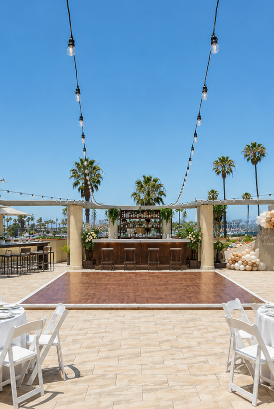 Rooftop wedding setup with white tables, blue seating, string lights, umbrellas, and palm trees under a clear blue sky.