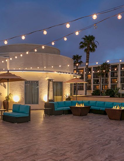 A stylish rooftop lounge with teal sofas, string lights, palm trees, and a modern circular bar at dusk, creating a cozy outdoor vibe.
