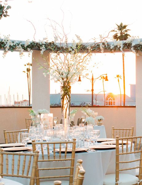 An outdoor wedding reception on a terrace with round tables, white linens, gold chairs, and flowers with a sunset backdrop.