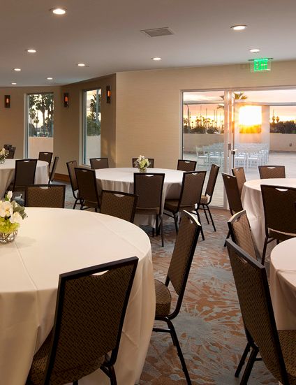 A banquet hall with round tables, chairs, white linens, flower centerpieces, and big windows showing a sunset outside.