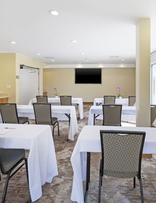 Bright conference room with set tables, chairs, white tablecloths, orchids, and a wall TV.