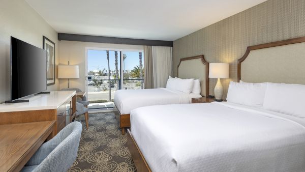 Bright hotel room with two beds, bedside lamps, patterned carpet, a flat-screen TV, and big windows opening to palm views.