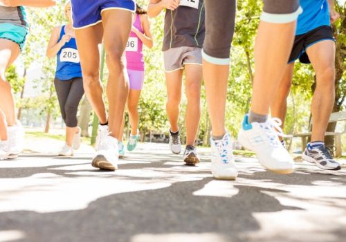 A group of runners mid-race on a sunny path, legs and shoes in motion, numbers pinned to shorts, trees in the background.