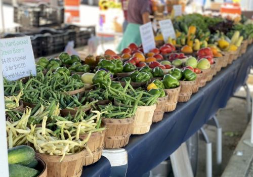 A market stall displays baskets of fresh garden greens and vegetables, neatly lined up on a blue-clothed table with signs and shoppers in the background.