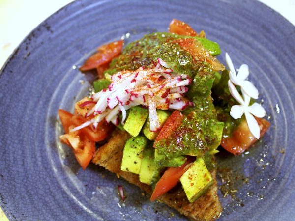 A colorful veggie toast or salad bowl with chopped avocado, tomatoes, cucumber, radish, greens, topped with bright green herb sauce and white flower garnish, ending with flair.