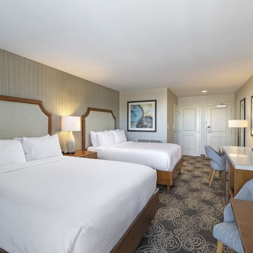 A modern hotel room with two beds, lamps, a desk and flat-screen TV, plus patterned carpet and framed wall art.