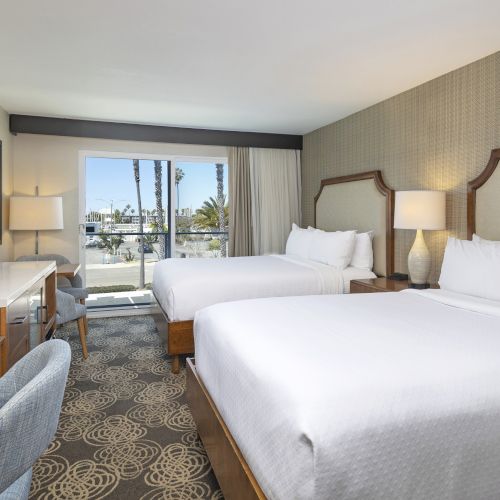 A modern hotel room with two beds, bedside lamps, a flat-screen TV, and large windows showing palm trees outside.
