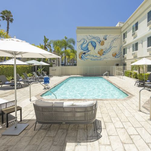 A sunny hotel courtyard with a swimming pool, lounge chairs under white umbrellas, palm trees, and a mural on the building.