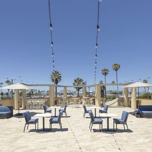 Sunny outdoor patio with lounge seating, small tables under white umbrellas, hanging string lights, and palm trees in the background.