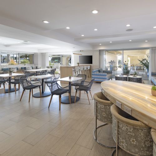 Bright modern lounge/patio with outdoor furniture, high-top tables, bar seating, a TV, and indoor plants under recessed ceiling lights.