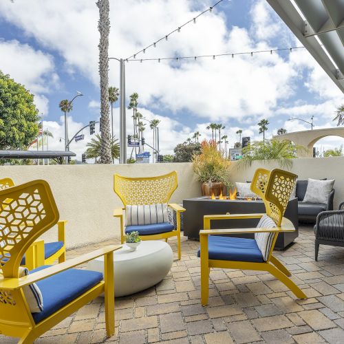 Outdoor patio with bright yellow chairs, a fire pit, string lights overhead, and palm trees under a cloudy blue sky.