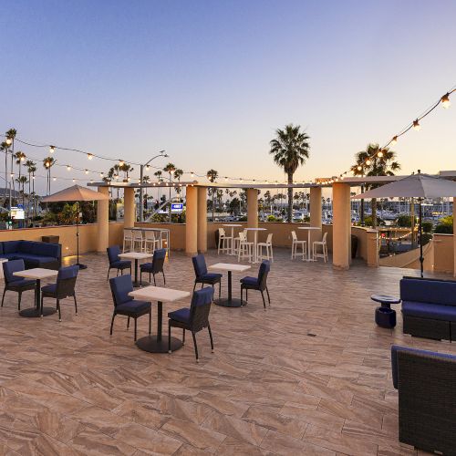 An outdoor patio with string lights, lounge seating and tables, palm trees, and a harbor view at sunset, creating a relaxed evening.