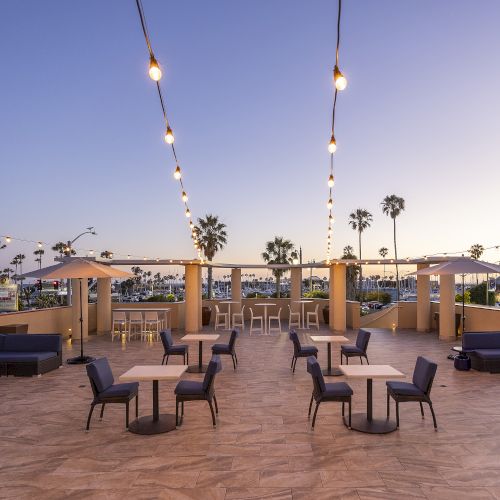 Rooftop patio lounge with string lights, tables and sofas, palm trees in the distance at sunset.