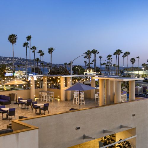 An oceanfront hotel patio at dusk with string lights, lounge chairs, palm trees, and a seaside promenade stretching into the distance.