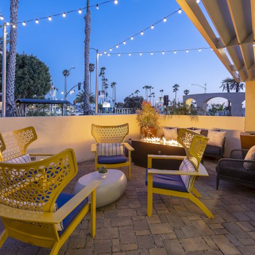 A sunny evening patio with yellow chairs, a cozy fire pit, string lights overhead, and a palm-lined walkway beyond.