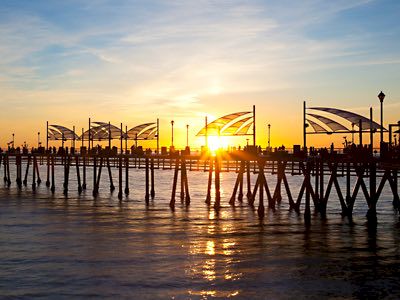 Sunset over a wooden pier with stilts, flags and curved canopies, silhouettes of people and structures against a golden sky, calm water below.