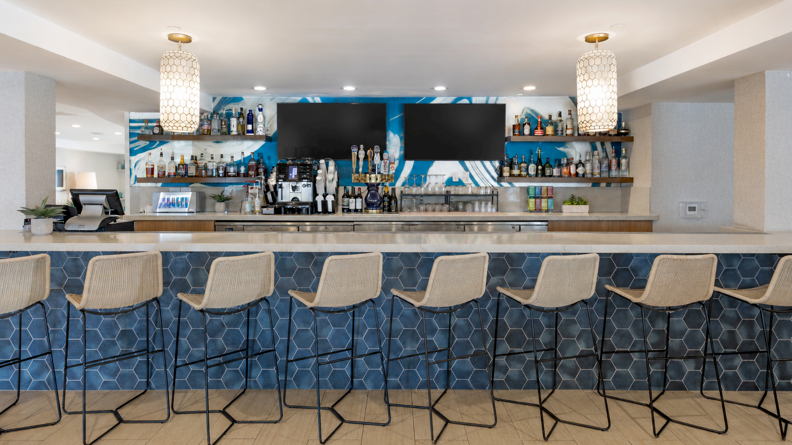 A modern bar with a bottle-lined back wall, two TVs, pendant lights, and six wicker stools along a blue tiled counter.
