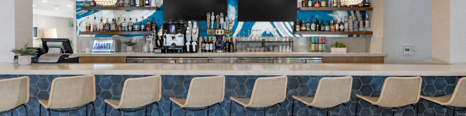 A modern bar with a bottle-lined back wall, two TVs, pendant lights, and six wicker stools along a blue tiled counter.
