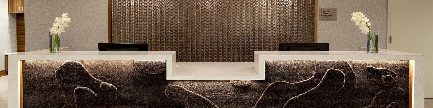 A sleek hotel reception with a dark wood front, glowing edge lighting, decorative wall panel, modern pendant lights, and fresh vases of white orchids.