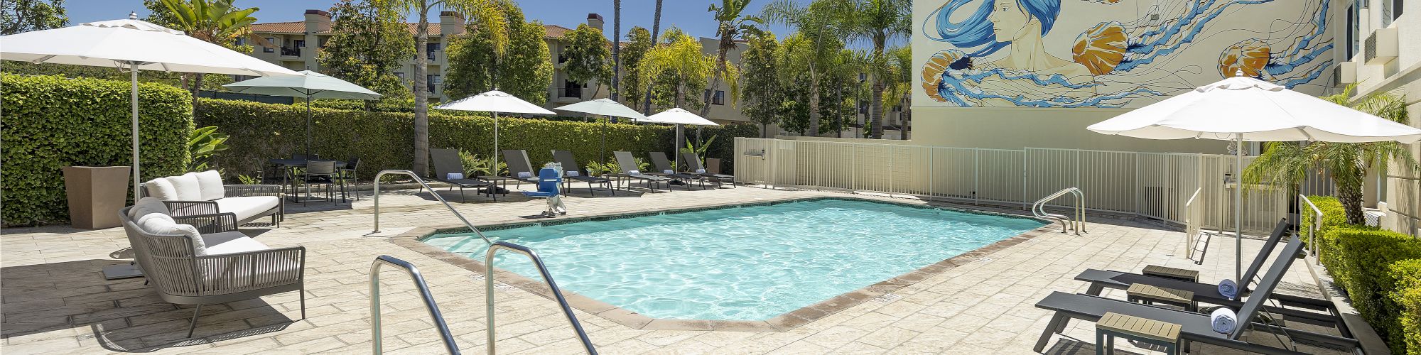 A sunny hotel courtyard pool with lounge chairs and a colorful mural of a mermaid on the building wall.
