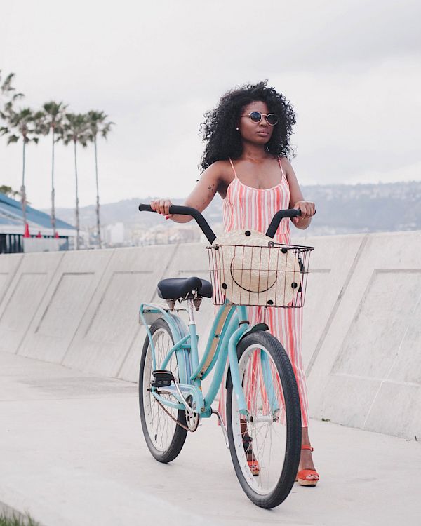 A woman with curly hair in a pink dress stands with a pastel bicycle by a sunny boardwalk, palm trees lining the path near the ocean.