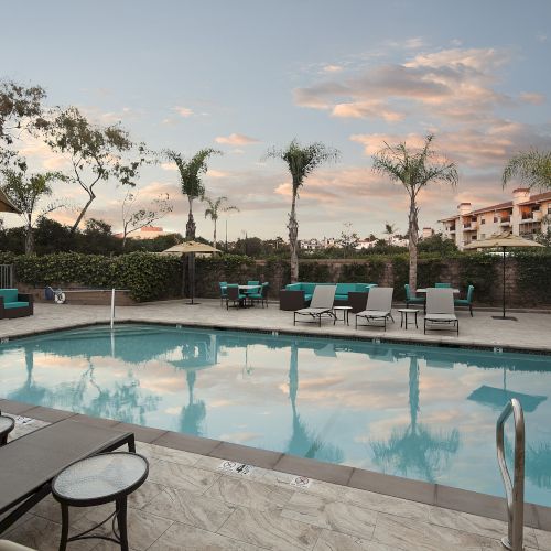 A resort-style pool area with sun loungers, umbrellas, and palm trees, surrounded by a low fence and nearby buildings at sunset. End.