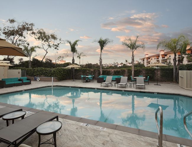 A resort-style pool area with sun loungers, umbrellas, and palm trees, surrounded by a low fence and nearby buildings at sunset. End.
