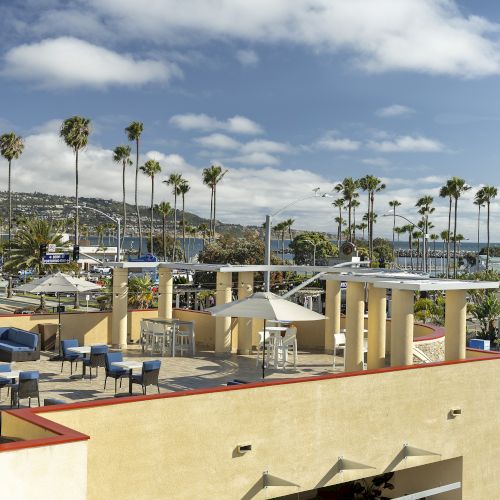Sunny resort terrace with palm trees, blue ocean view, and caf&eacute; seating under a cloudy sky.