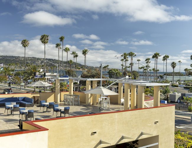 Sunny resort terrace with palm trees, blue ocean view, and caf&eacute; seating under a cloudy sky.