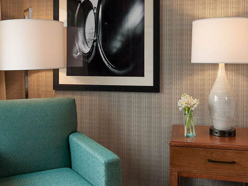 A modern hotel room with a teal chair, wooden nightstand, lamps, and a neatly made bed by a window view of palm trees.