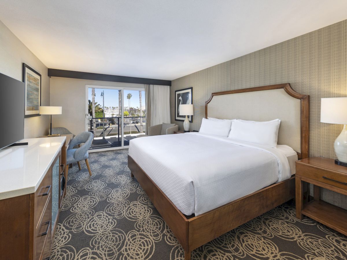 A bright hotel bedroom with a large bed, bedside lamps, patterned carpet, and a window showing palm trees.